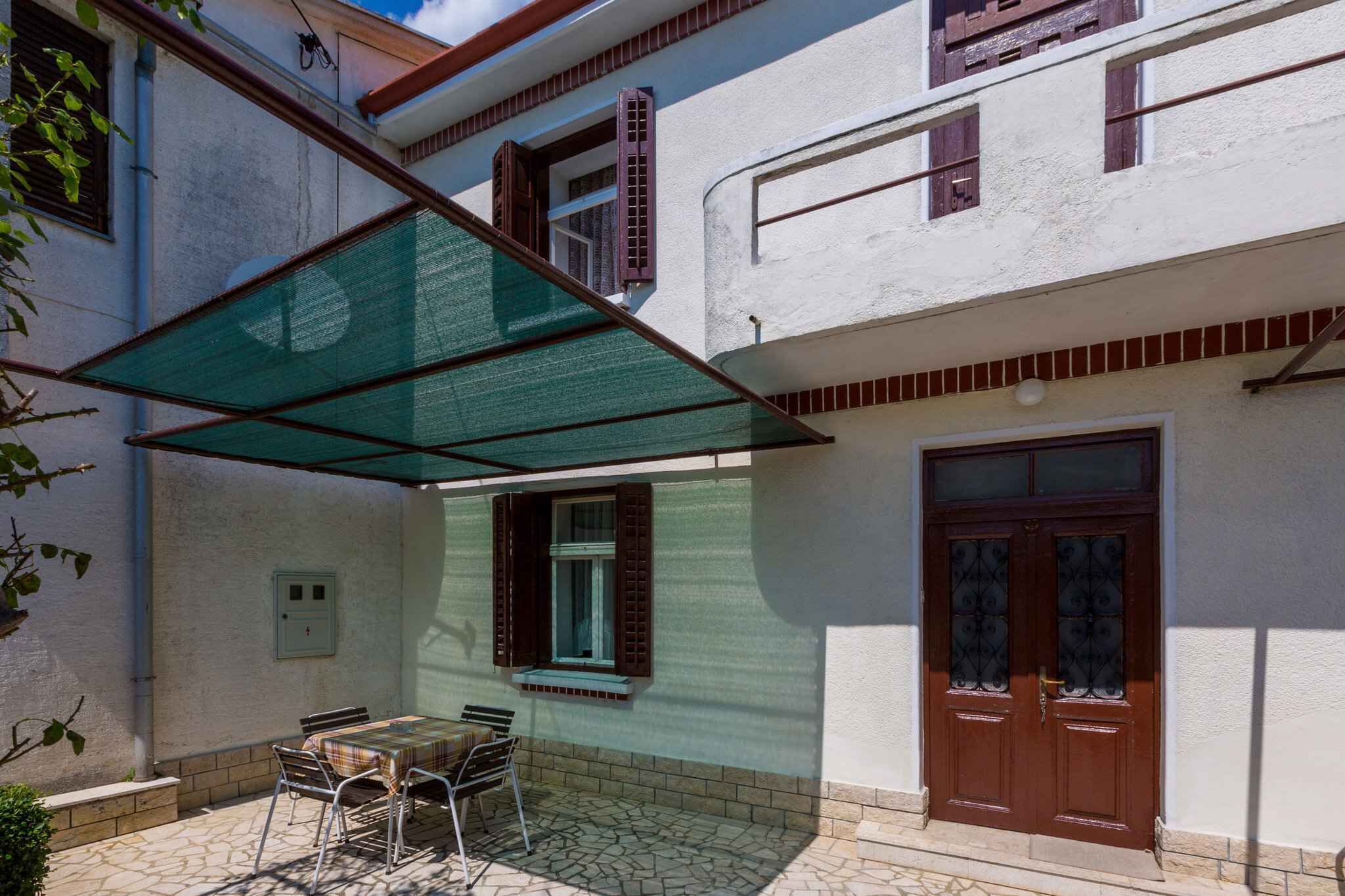 A covered outdoor patio with a table and chairs, a door, and a window with shutters.