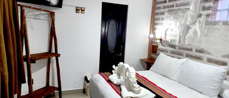 A hotel room with a bed, a TV mounted on the wall, a wooden chair, a green rug, and a decorative wall piece.