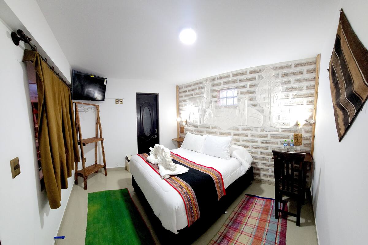 A bedroom with a bed, a green rug, a plaid rug, a wooden ladder, a black door, a TV, and a brick wall.