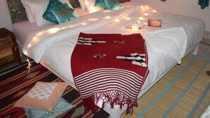 Un lit avec du linge de lit blanc, plusieurs oreillers et une couverture rouge. On y voit des guirlandes lumineuses, une petite table avec une bougie et une tête de lit décorative.