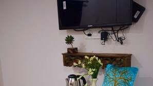 Une petite pièce agréable et chaleureuse, avec un meuble en bois, un téléviseur fixé au mur et une petite table sur laquelle se trouvent un vase et une bouilloire.