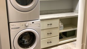 A compact laundry room with two stacked front-loading washing machines, a countertop with a bottle of detergent, and a wooden floor. - Park Avenue Loft overlooking Parkerson Avenue (Crowley)