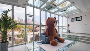 En rummelig stue med et stort glasbord, en blød brun bamse og udsigt til en traditionel bygning gennem store vinduer.