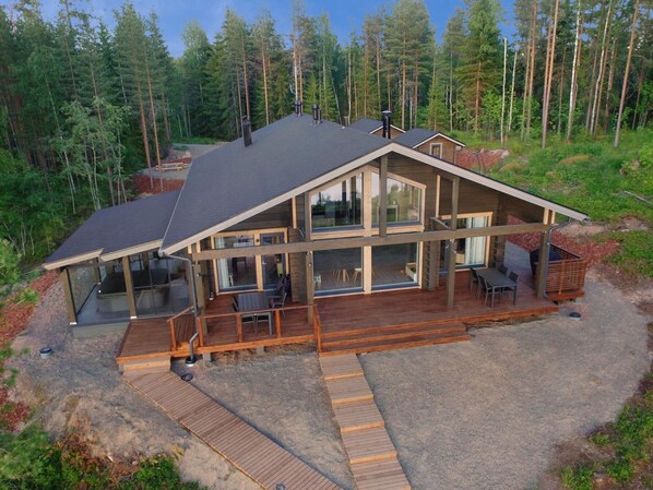Une maison moderne à deux étages, dotée de grandes fenêtres, d’une terrasse en bois et entourée d’une forêt.