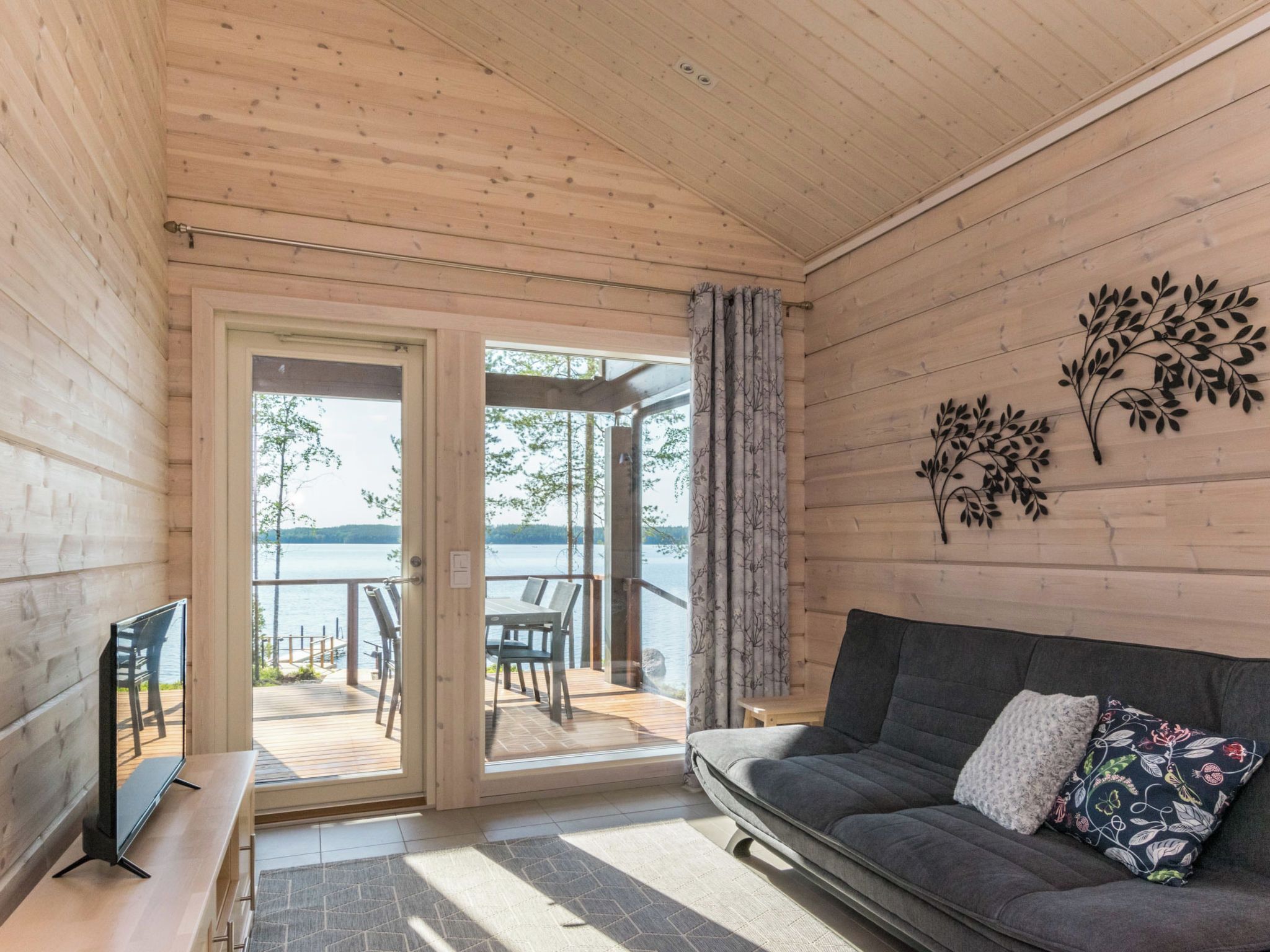 Un interior de madera con un sofá, un televisor y vista a un lago a través de una puerta de cristal.