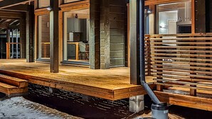 Una terraza de madera con barandilla metálica, un área de cocina visible con armarios y un lavabo, y una ventana con vista al interior.