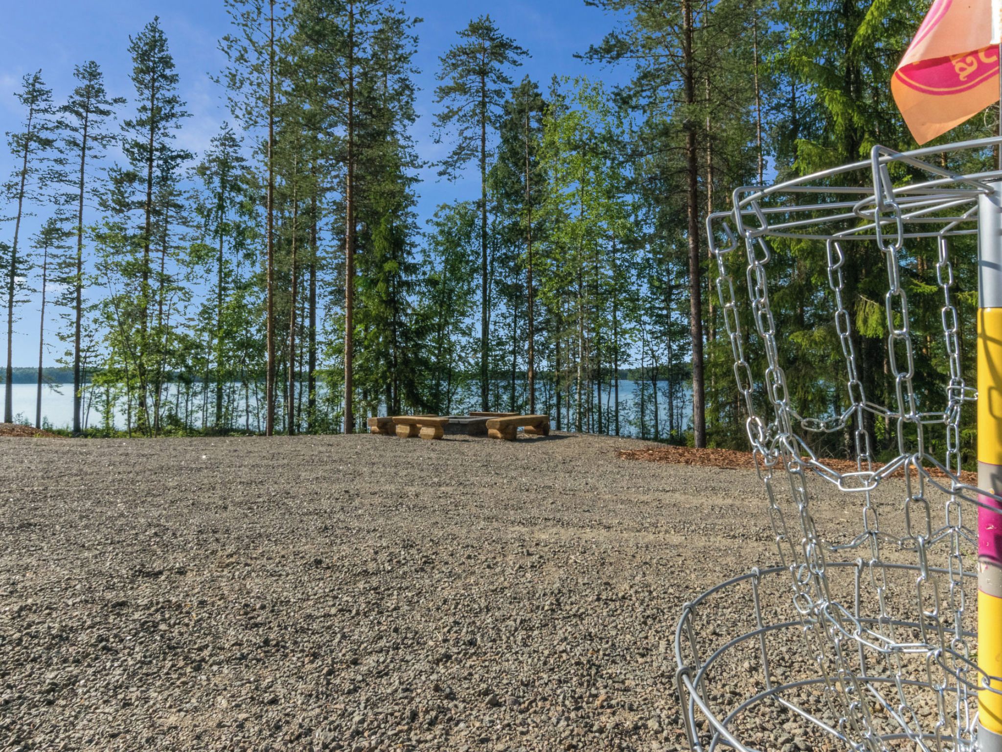 En grusmark med et metalmål, omgivet af høje træer og et vandareal i baggrunden.