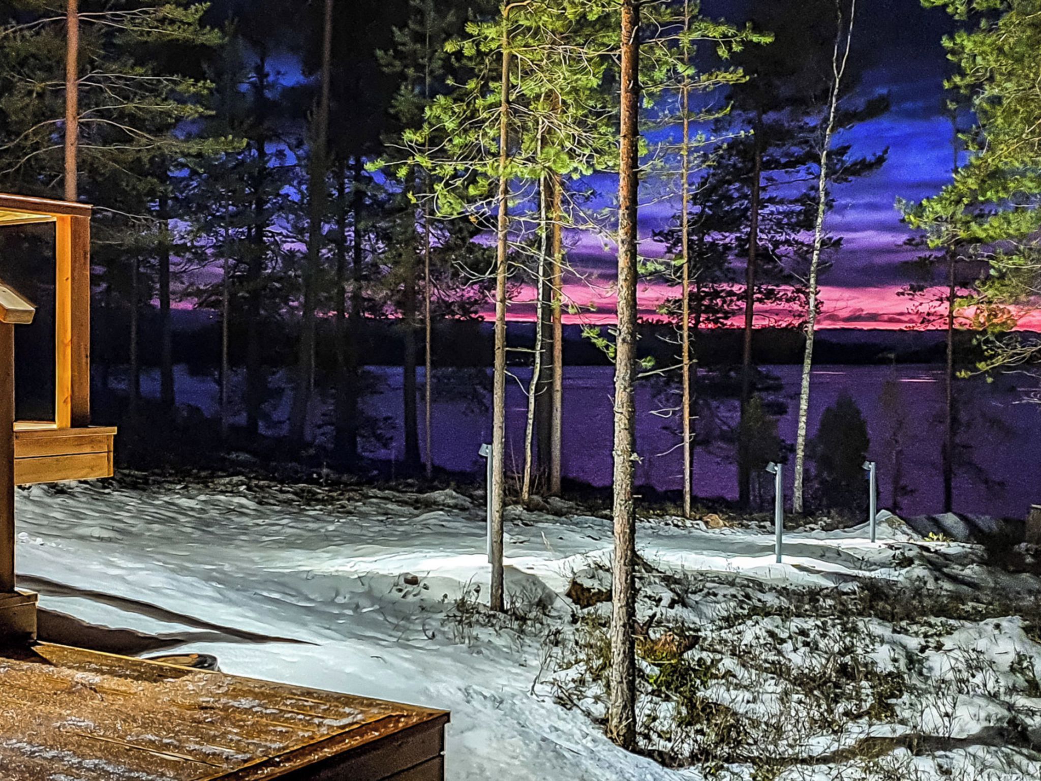 Eine Holzhütte mit einer Holzdiele, umgeben von verschneitem Gelände und Bäumen, mit Blick auf einen See und einen Sonnenuntergang.