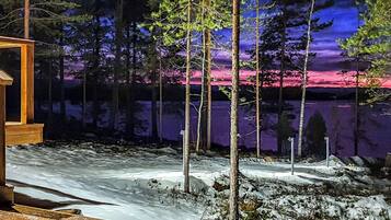 Eine Holzhütte mit einer Holzdiele, umgeben von verschneitem Gelände und Bäumen, mit Blick auf einen See und einen Sonnenuntergang.