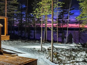 A cabin with a wooden deck, surrounded by snow-covered ground and trees, with a view of a lake and a sunset.