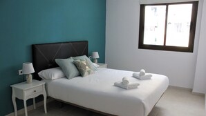 Una cama bien hecha con sábanas blancas y dos almohadas, una mesita de noche con lámpara y una ventana con vistas a edificios.