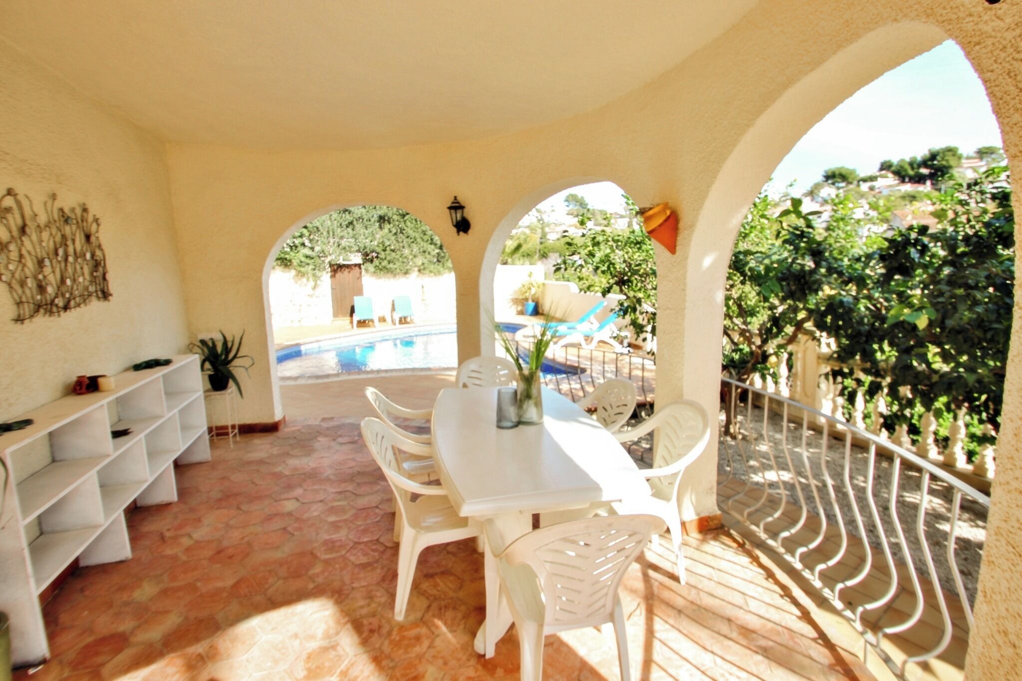 A sunlit patio with a white table and chairs, a potted plant, and a view of a swimming pool and greenery.