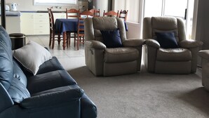 A spacious living room with a blue sofa, two beige recliners with blue cushions, a wooden ceiling, and a dining area in the background.