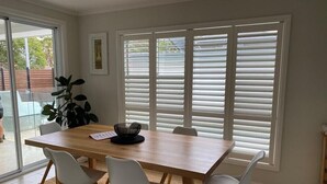 Een eetgedeelte met een houten tafel, vier witte stoelen en een groot raam met witte luxaflex.