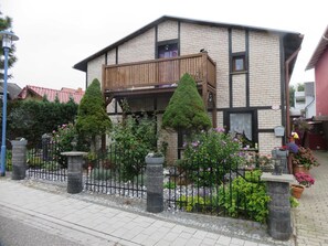 A two-story house with a balcony, surrounded by a stone fence and various plants.