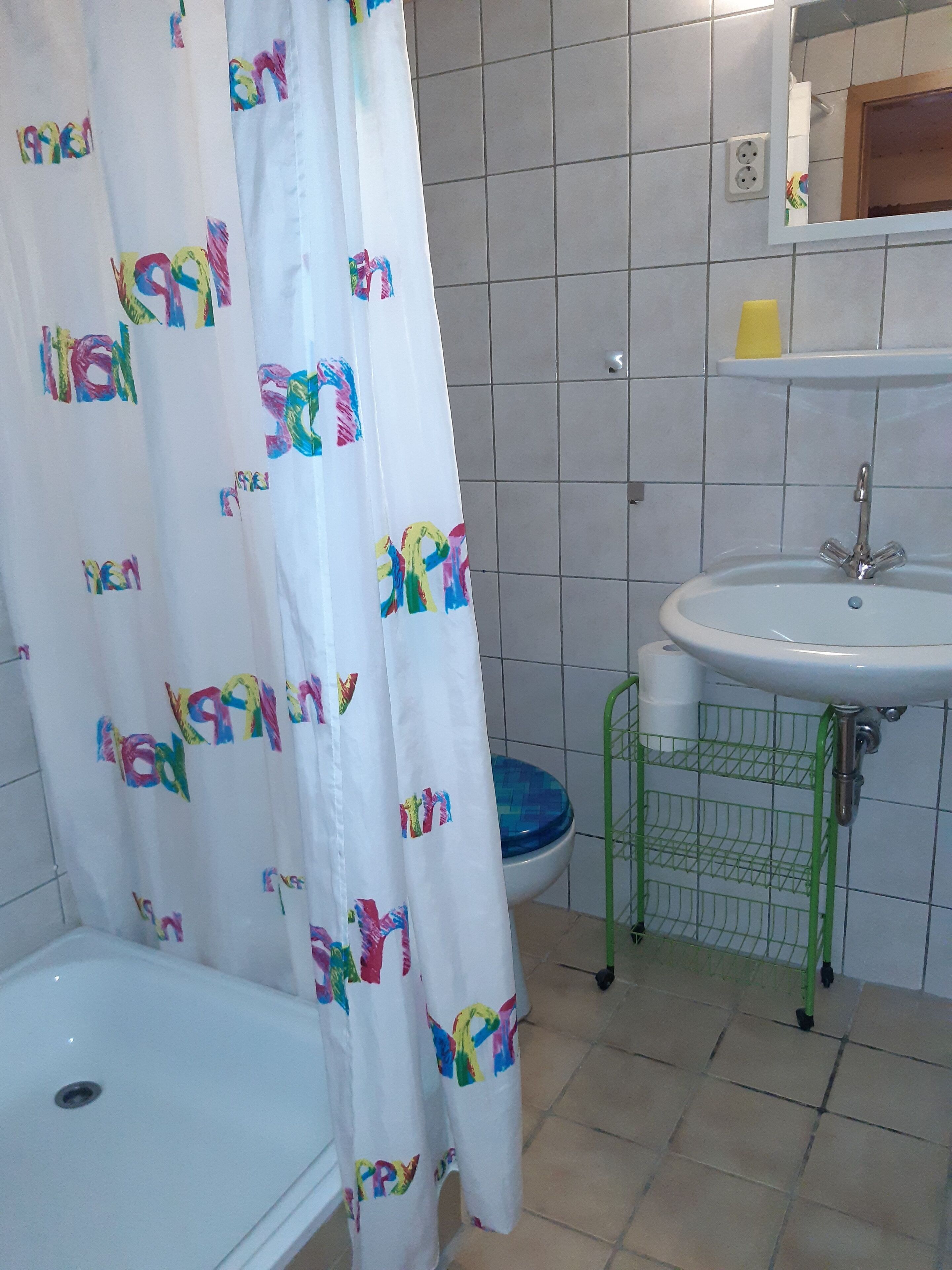 Ein Badezimmer mit einem weißen Waschbecken, einem grünen Metallregal, einer blauen Toilette und einem weißen Duschvorhang mit farbigem Text.