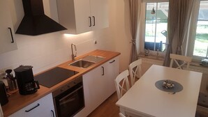 Eine kleine Küche mit Spüle, Herd und weißen Schränken. Ein Esstisch mit Stühlen und ein Fenster mit Vorhängen sind ebenfalls vorhanden.
