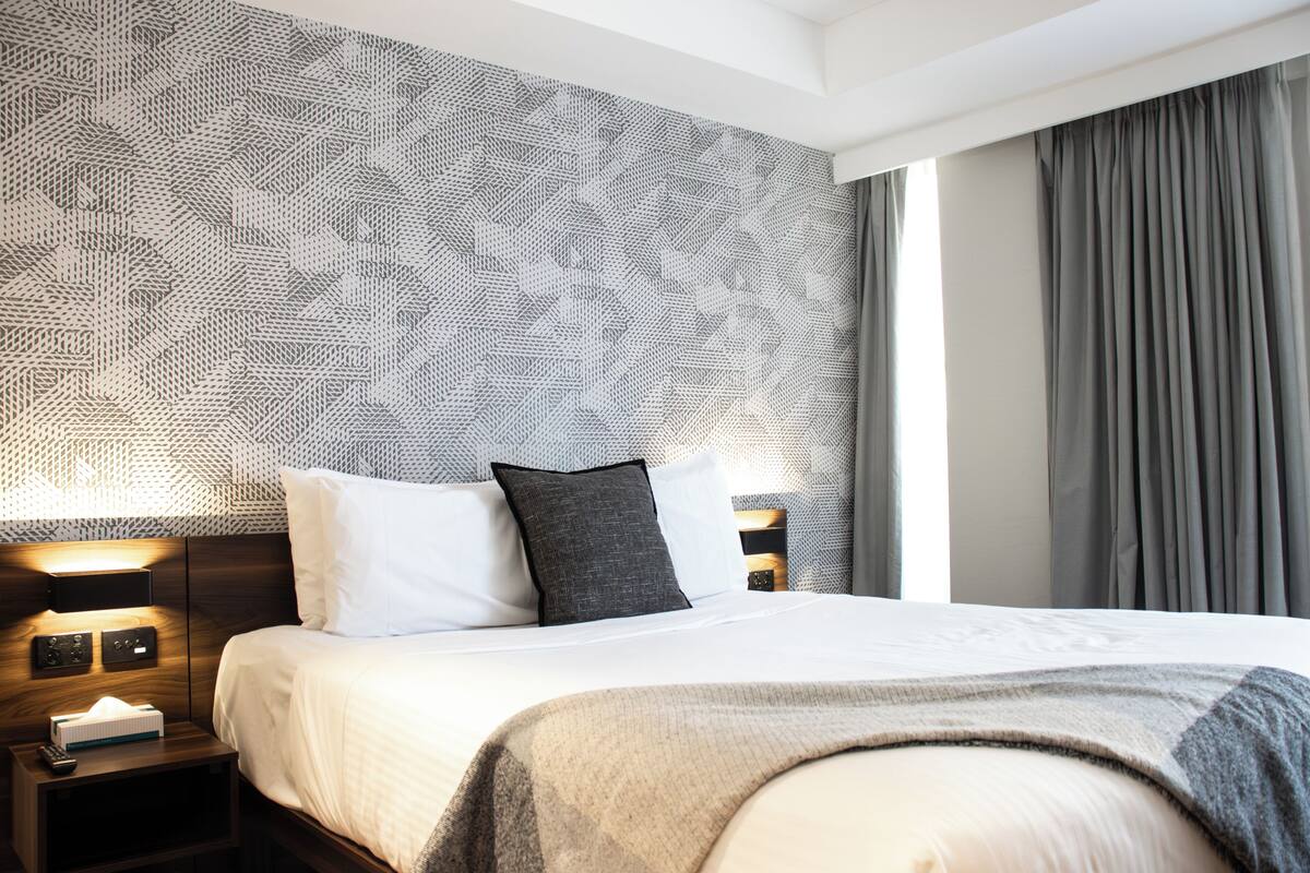 A hotel room with a patterned wall, a bed with white and gray bedding, and a nightstand with a remote control.