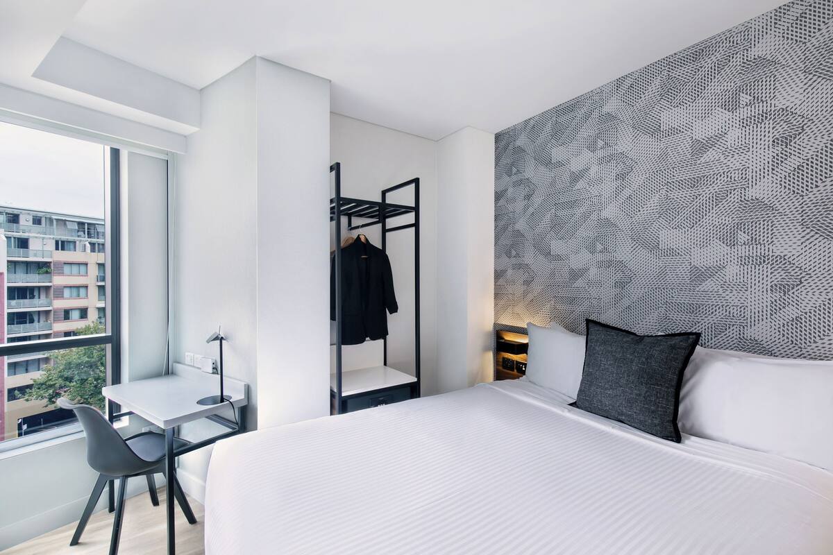 A modern hotel room with a large bed, a desk, and a chair. There is a wardrobe with a mirror and a hanging rack. The room has a large window and a patterned wall.
