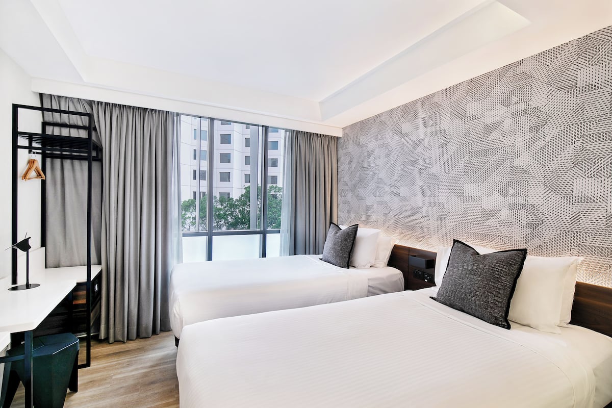 A hotel room with two beds, a desk, and a view of buildings through large windows.