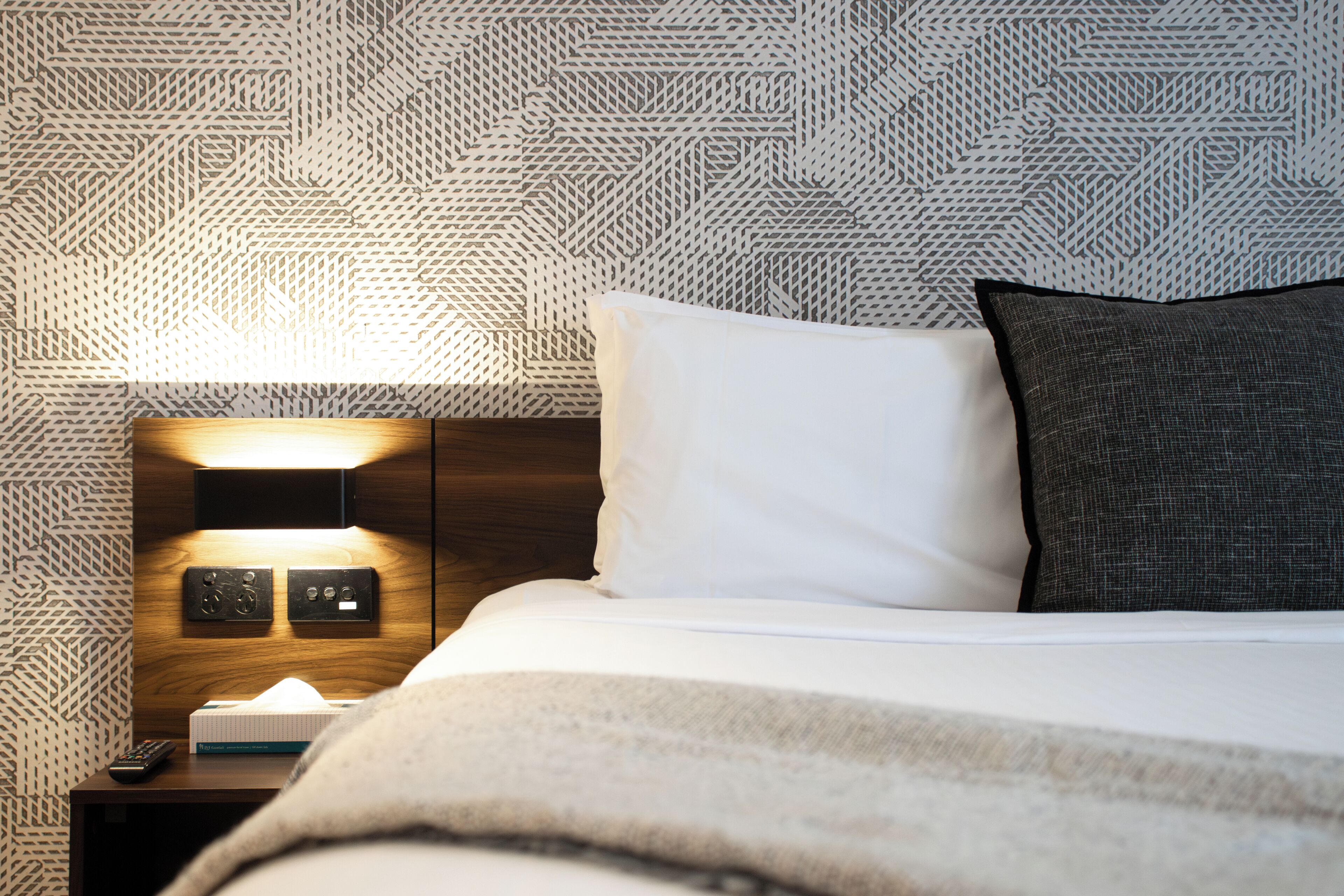 Una cama bien hecha con almohadas blancas y oscuras, una mesita de noche con control remoto, una lámpara y un reloj de pared.