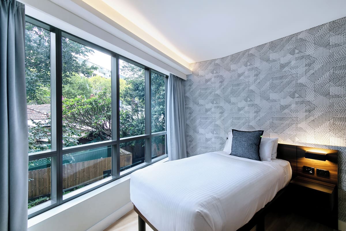 A bedroom with a large window, a bed with white bedding, a patterned wall, and a nightstand with a lamp.