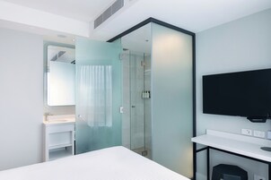 Une chambre d’hôtel moderne comprenant un lit, une salle de bain avec une douche à parois de verre, un téléviseur fixé au mur et une table de chevet blanche.