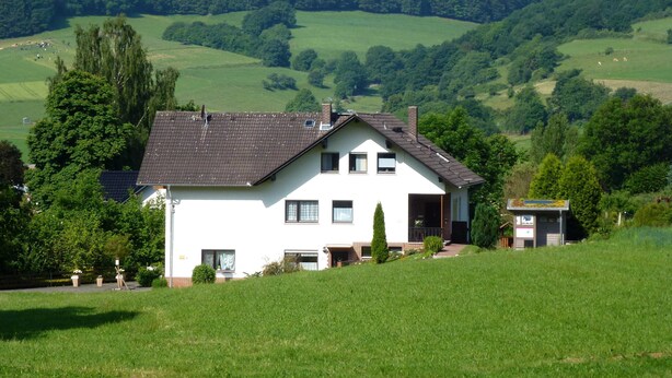 Une maison à deux étages, dotée d’un toit en pente, entourée de champs verdoyants et d’arbres.