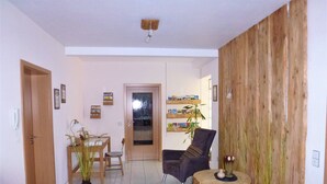 Un salon avec un plancher en bois, une table basse ronde, un canapé et une table à manger avec des chaises. On y trouve une porte avec un panneau de verre, une étagère avec des livres et une plante.