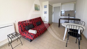 A living room with a red patterned sofa, a small side table, a white lamp, and a dining area with a white table and black chairs. - STUDIO AVEC PKG, LES CRIQUES DE LA CORNICHE (Sète)