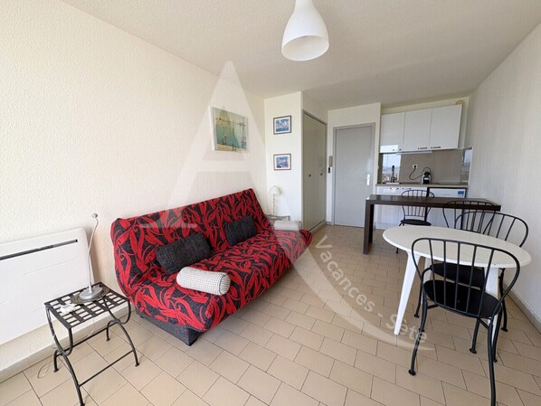 A living room with a red patterned sofa, a small side table, a white lamp, and a dining area with a white table and black chairs. - STUDIO AVEC PKG, LES CRIQUES DE LA CORNICHE (Sète)