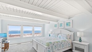 A bedroom with a large bed, two bedside tables, a desk, and a chair. There are three windows with a view of the beach. - Ocean Colony 8 by Seaside Vacations & Sales (Ocean City)