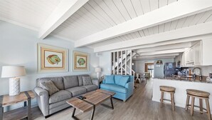 A spacious living area with a grey sofa, blue armchair, and a wooden coffee table. A kitchen with white cabinets and a bar area is visible in the background. - Ocean Colony 8 by Seaside Vacations & Sales (Ocean City)