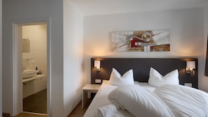 Une chambre d’hôtel avec un grand lit, deux tables de chevet, une œuvre d’art accrochée au mur et une salle de bain visible à travers une porte ouverte.