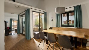 Un espace repas moderne avec une table et des chaises en bois, de grandes fenêtres avec des rideaux et une vue sur l’extérieur.
