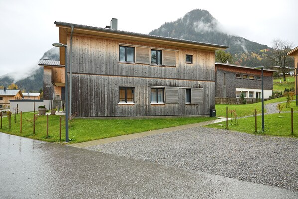 A two-story wooden house with a gravel driveway and a mountain backdrop.