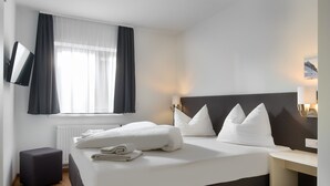 Une chambre d’hôtel moderne dotée d’un grand lit, d’un téléviseur à écran plat fixé au mur, d’une table de chevet avec une lampe et offrant une vue sur l’extérieur à travers des rideaux légers.