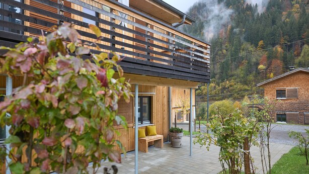 Un bâtiment moderne à l’extérieur en bois, doté d’un espace extérieur couvert avec des sièges et offrant une vue sur une montagne boisée.