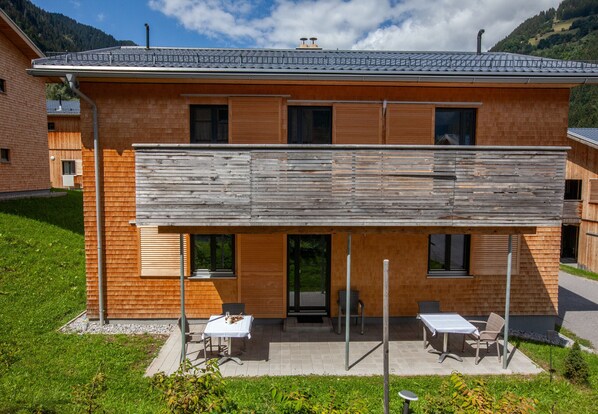 Un edificio moderno de dos plantas con fachada de madera, un área de estar exterior cubierta con mesas y sillas, y vistas a un paisaje montañoso.
