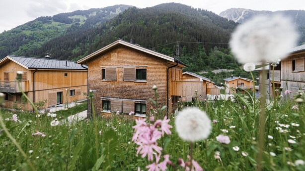 Ein hölzernes Haus mit geneigtem Dach, umgeben von üppiger Vegetation und Bergen im Hintergrund.