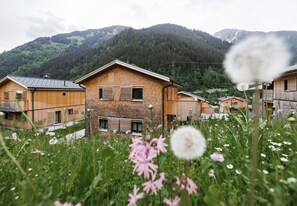 Una casa de madera con techo inclinado, rodeada de abundante vegetación y montañas al fondo.
