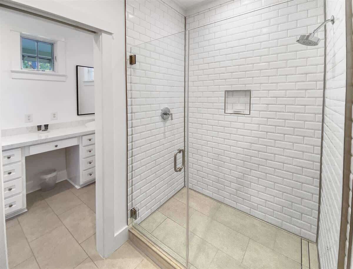 Ein Badezimmer mit weißen Ziegelwänden, einer Duschkabine aus Glas, einem weißen Waschtisch mit Schubladen und einem Fenster.