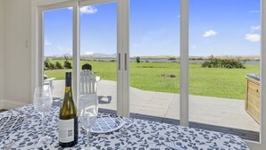 A dining area with a table set for two, a bottle of wine, and a glass. The table is covered with a patterned tablecloth. Large sliding glass doors lead to an outdoor patio with a view of a grassy field and a body of water.