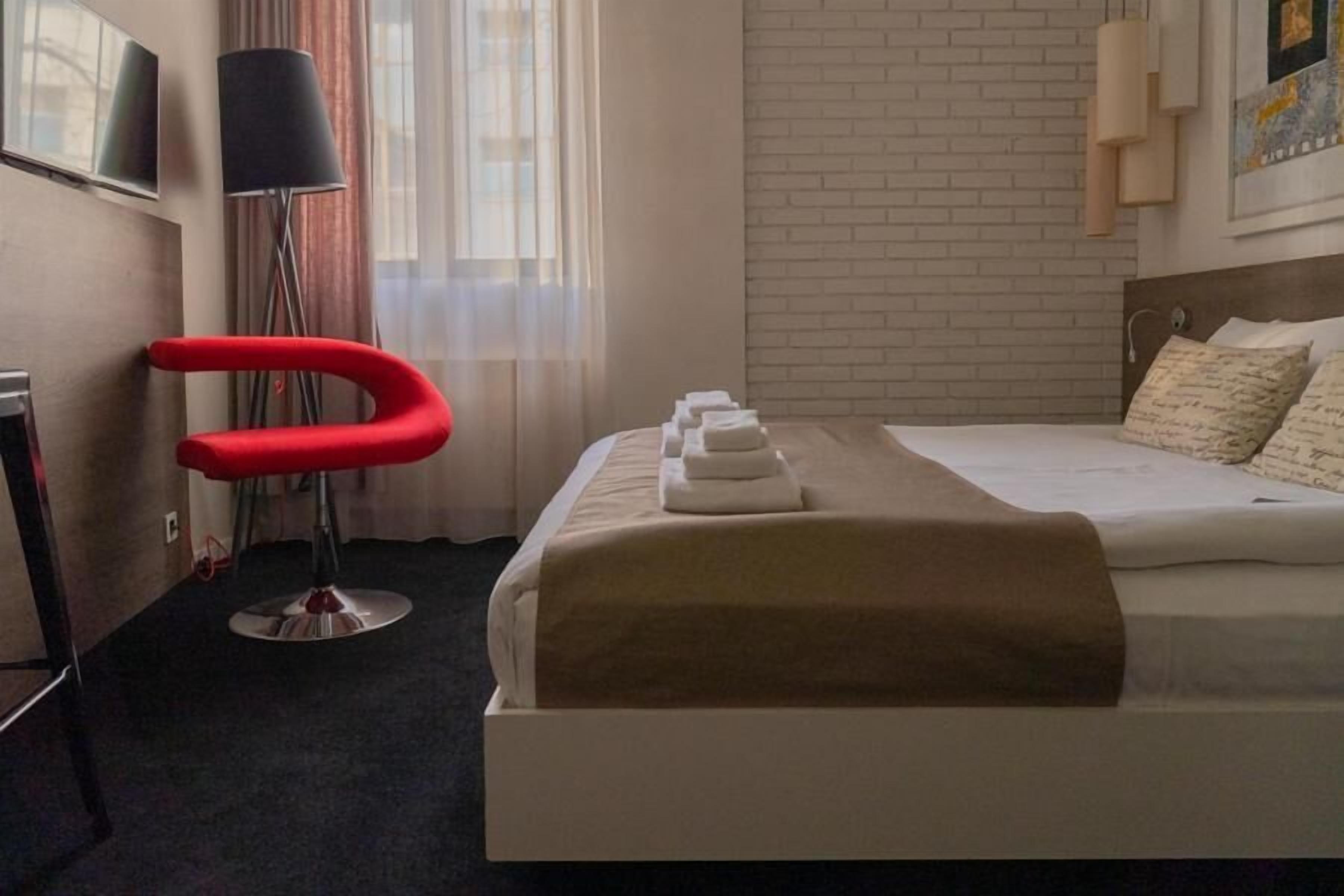 A hotel room with a bed, a red chair, a lamp, and a window with curtains.