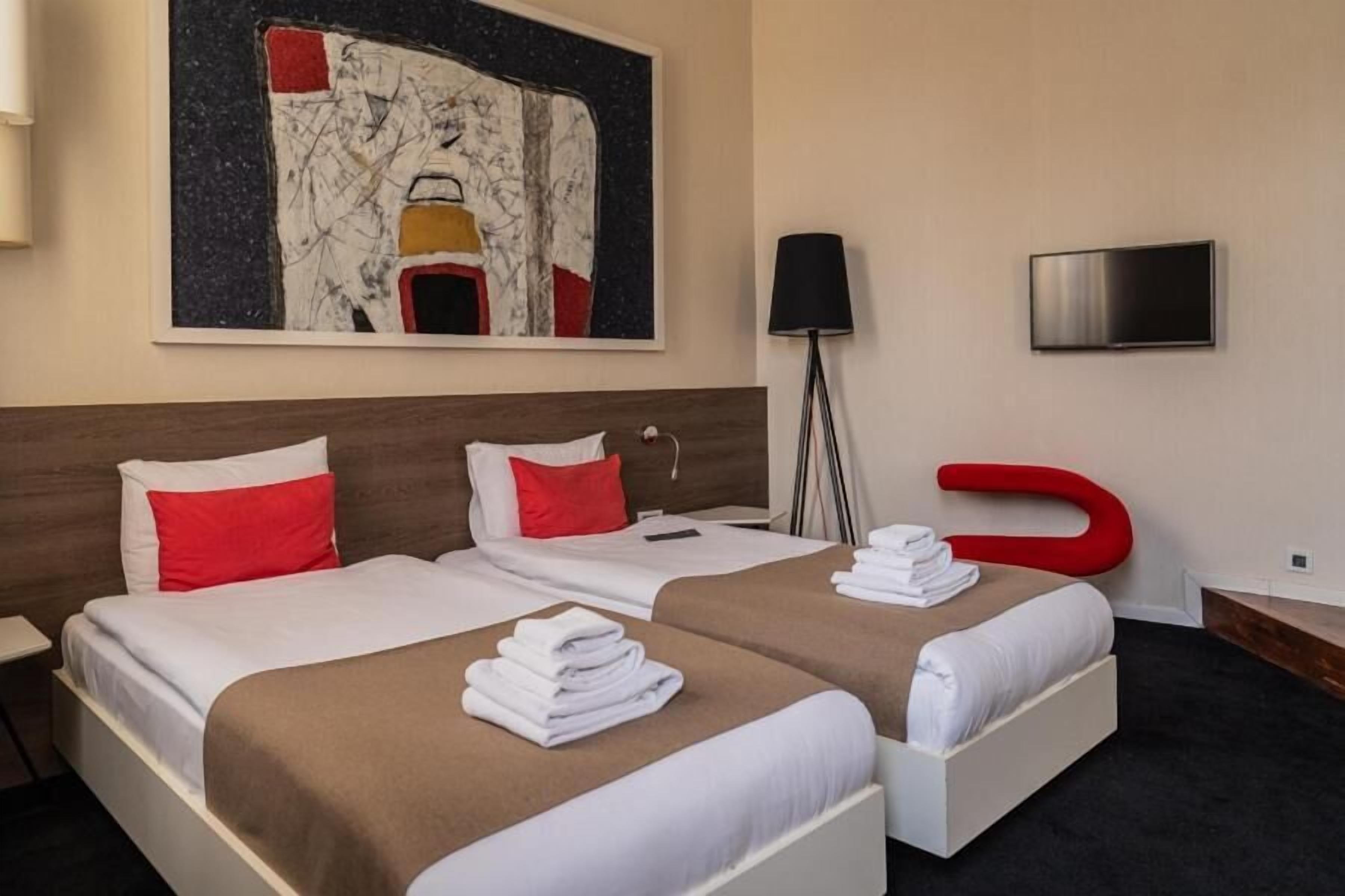 Ein Hotelzimmer mit zwei Betten, einem modernen abstrakten Gemälde, einem Flachbildfernseher und einem auffälligen roten Sessel.