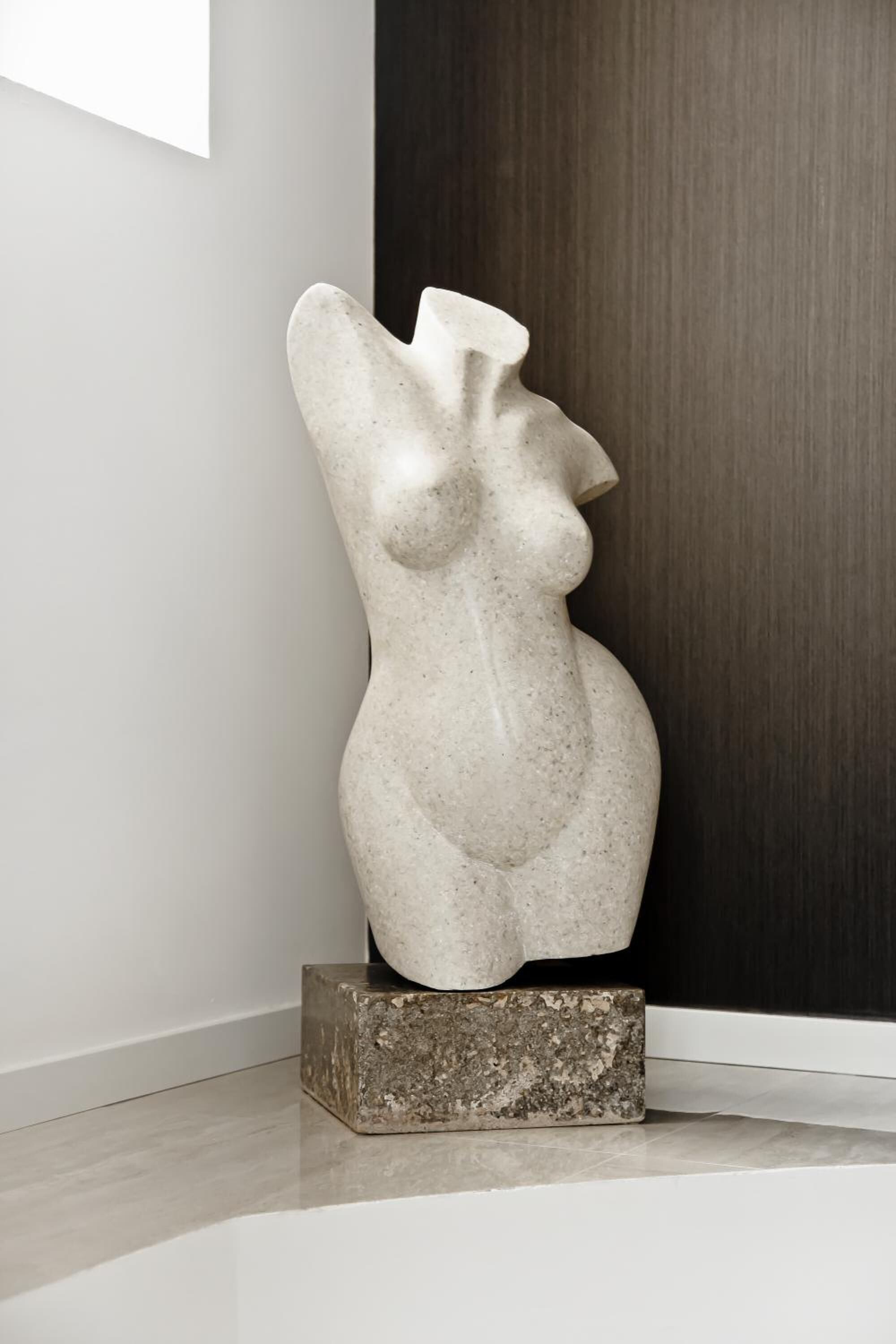 Eine Skulptur einer weiblichen Figur auf einem Sockel in einem Raum mit einer dunklen Holztür und einer weißen Wand.