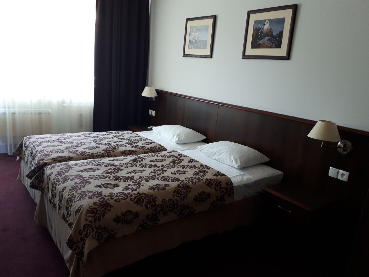 Een hotelkamer met twee bedden, donkere gordijnen, twee wandlampen en twee ingelijste foto's.