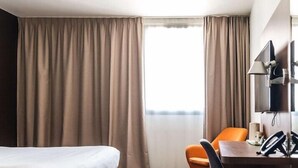 A hotel room with a bed, a desk with a chair, a television, and a window with curtains.