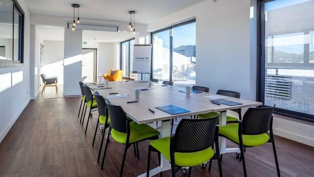 A modern conference room with a long table, green chairs, and large windows offering a view of the cityscape.
