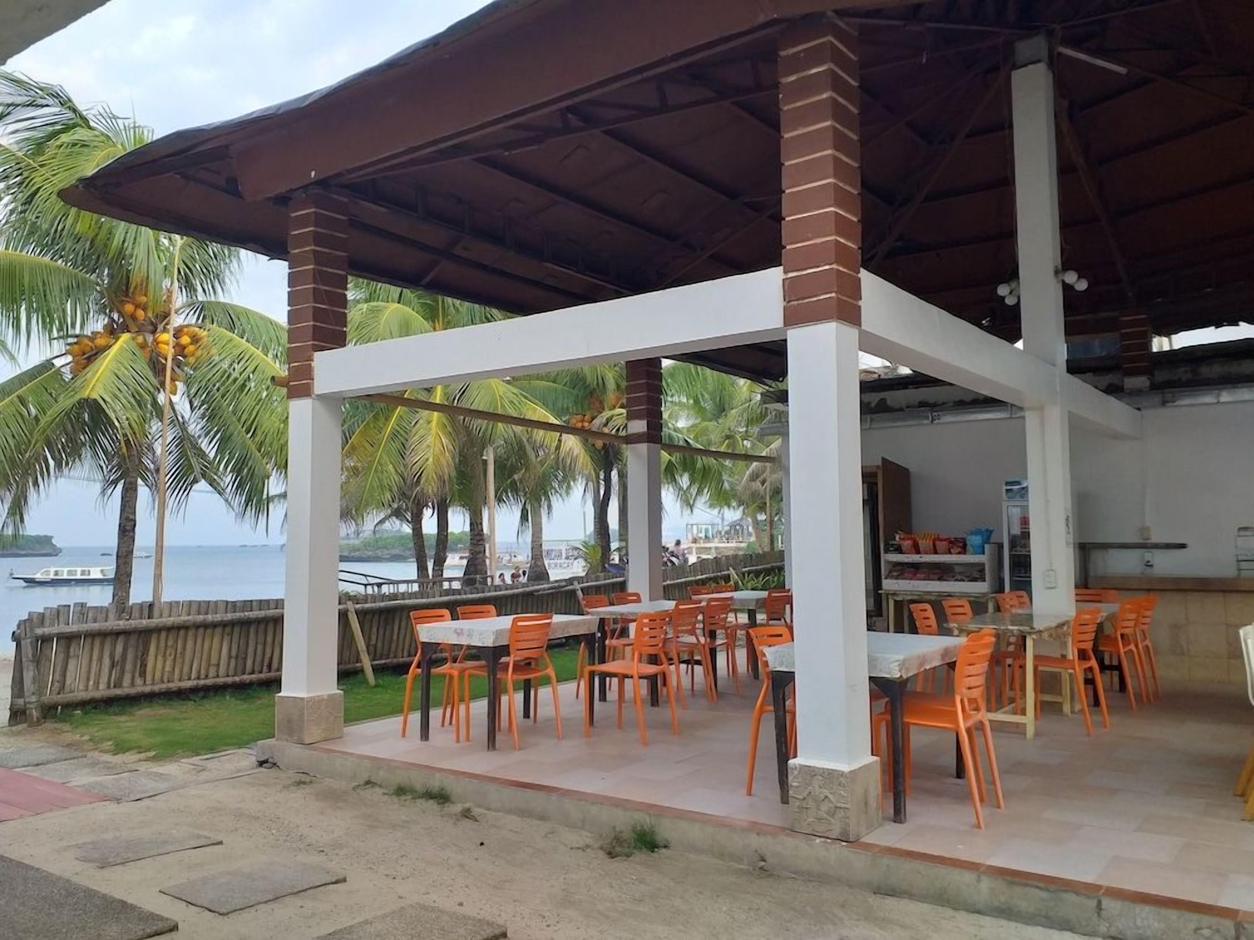 An outdoor seating area with tables and chairs, a covered structure, and a view of the sea and palm trees.